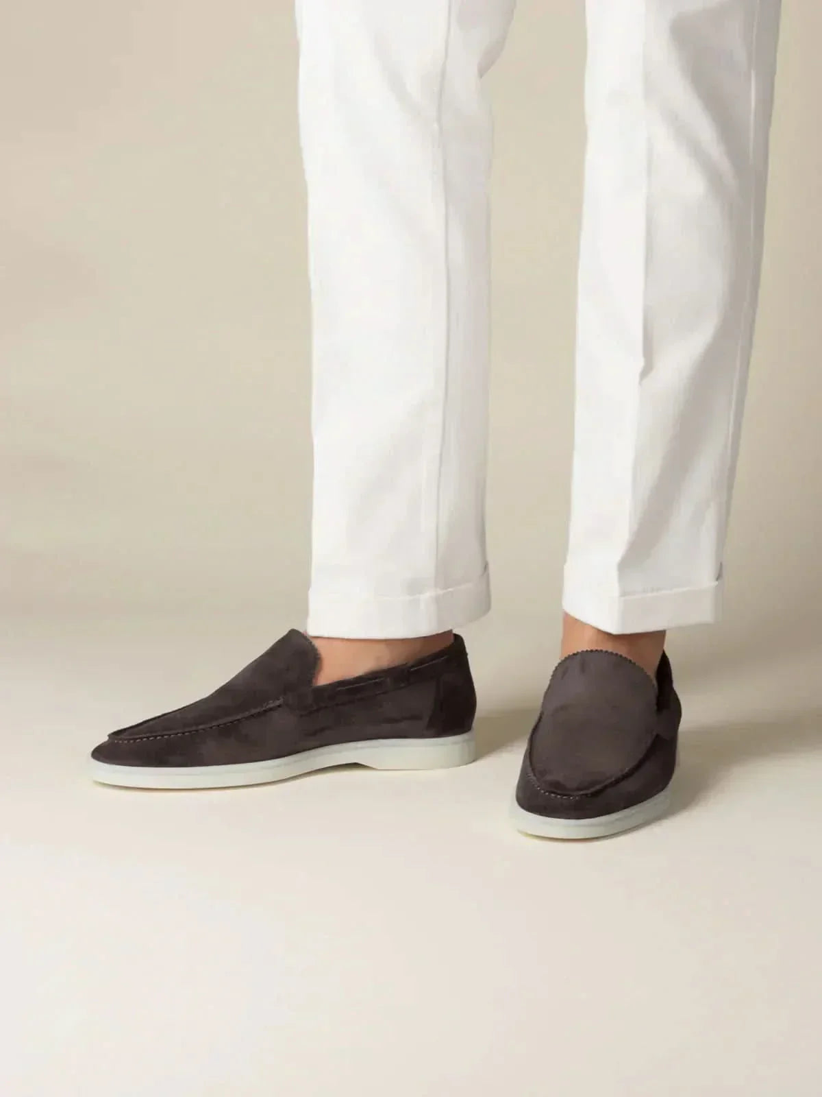 Mocasines de ante café oscuro, perfil lateral, con suela blanca.